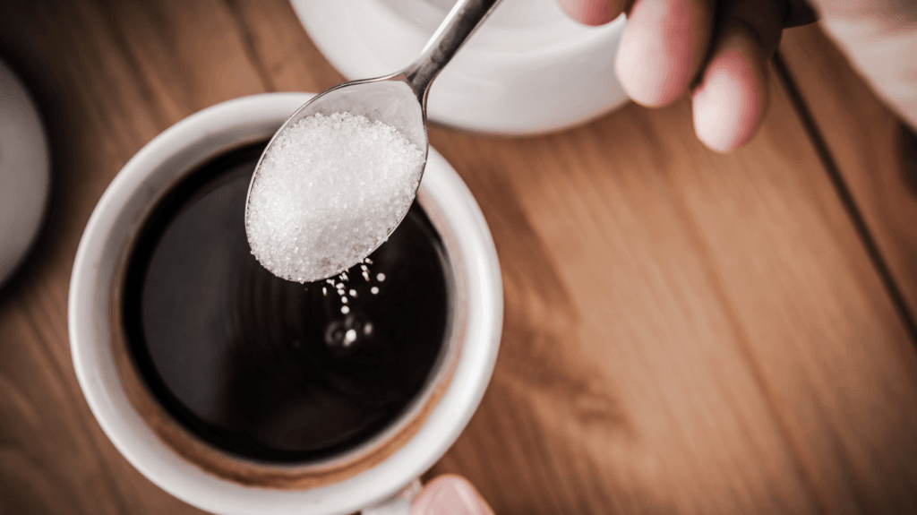 imagem de uma mão, segurando uma colher com pó branco, em cima de uma xícara de café puro. dando a entender que é açúcar.