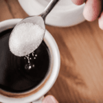 imagem de uma mão, segurando uma colher com pó branco, em cima de uma xícara de café puro. dando a entender que é açúcar.